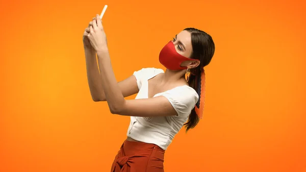 Woman in protective mask taking photo on smartphone isolated on orange - foto de stock
