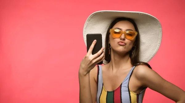 Jovem em maiô e óculos de sol segurando celular e lábios amuados isolados em rosa — Fotografia de Stock