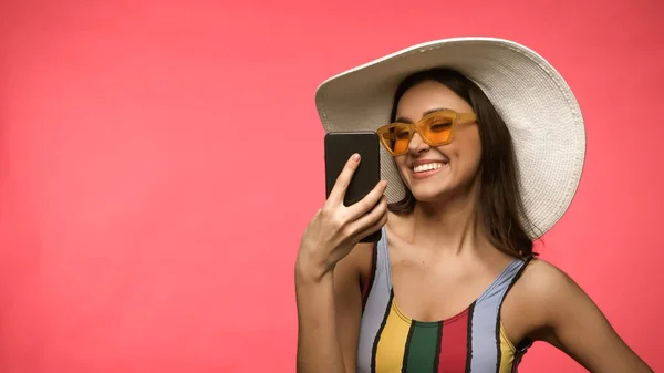 Mujer feliz en sombrero de sol y traje de baño con teléfono móvil aislado en rosa - foto de stock