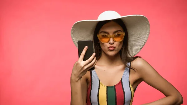 Woman in sun hat and swimsuit holding cellphone and pouting lips isolated on pink — Photo de stock