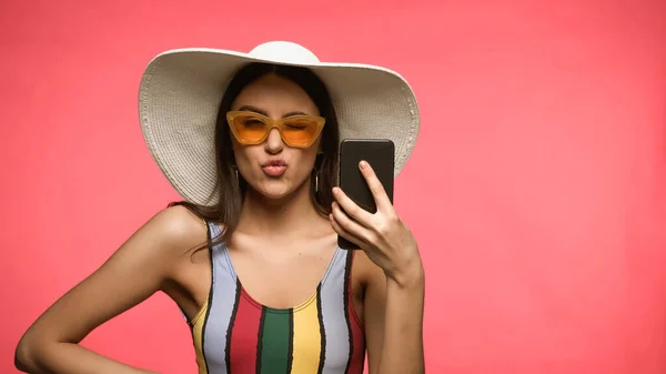 Hübsche Frau in Badeanzug und Sonnenhut hält Handy und macht Entengesicht isoliert auf rosa — Stockfoto