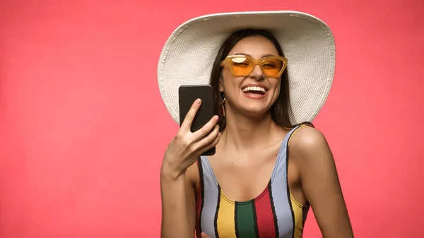 Mujer feliz en gafas de sol y sombrero de sol que sostiene el teléfono celular aislado en rosa - foto de stock