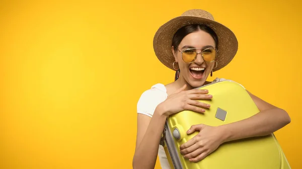 Excited woman in sun hat hugging suitcase isolated on yellow - foto de stock