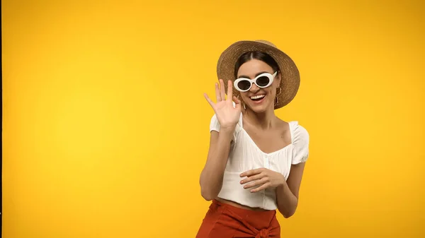 Усміхнена модель в сонцезахисних окулярах і солом'яному капелюсі махає рукою ізольовано на жовтому — стокове фото
