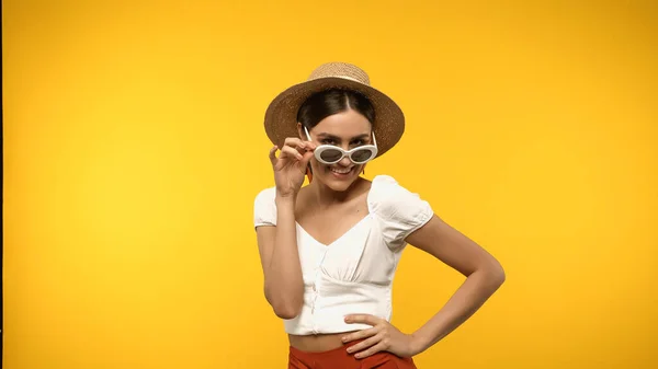 Mujer positiva en sombrero de paja y gafas de sol sosteniendo la mano en la cadera aislada en amarillo - foto de stock