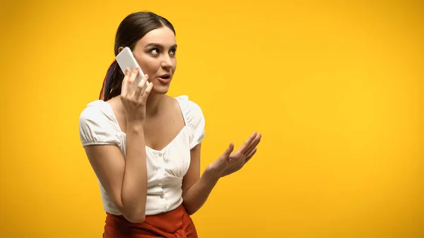 Pretty brunette woman talking on cellphone and gesturing isolated on yellow — Stock Photo