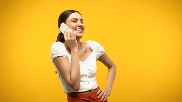 Positive brunette woman in blouse talking on cellphone isolated on yellow — стоковое фото
