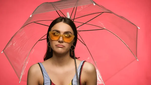 Disappointed woman in swimsuit and sunglasses looking at umbrella isolated on pink — стоковое фото