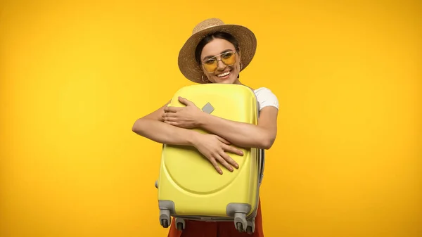 Joven viajero en gafas de sol abrazando maleta aislada en amarillo - foto de stock