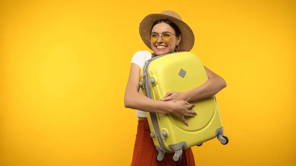Graziosa turista in cappello di paglia abbracciando valigia isolata su giallo — Foto stock