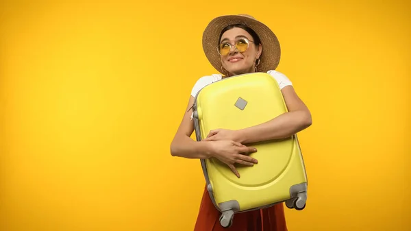 Smiling tourist in sunglasses holding baggage isolated on yellow — стоковое фото