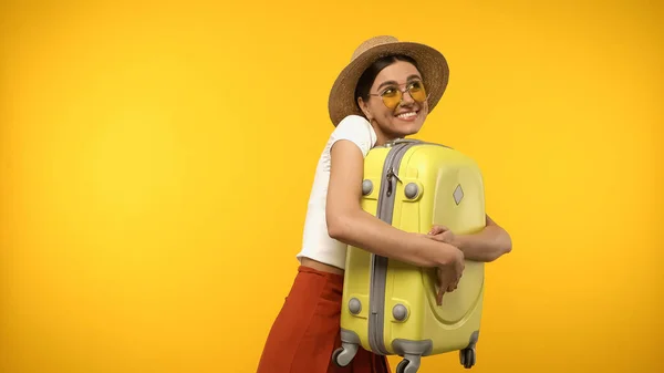 Viajero en gafas de sol y sombrero de paja abrazando maleta aislada en amarillo - foto de stock