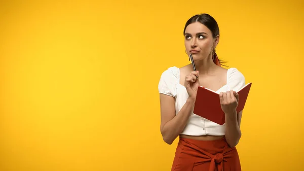 Pensive brunette woman holding pen and notebook isolated on yellow — Foto stock