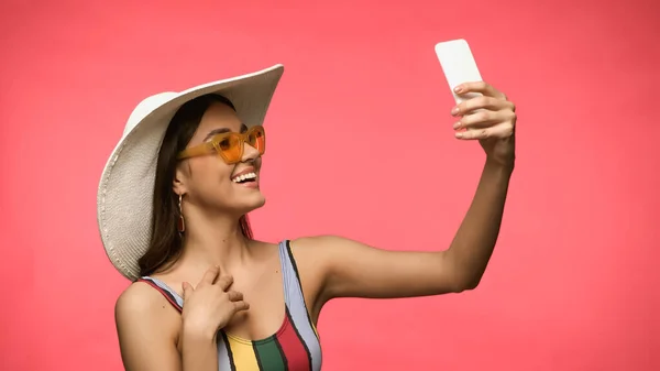 Mujer sonriente en traje de baño y gafas de sol tomando selfie en el teléfono inteligente aislado en rosa - foto de stock