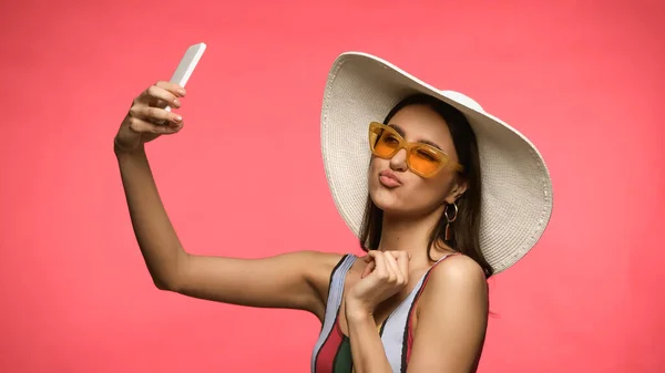 Bruna donna in cappello da sole e occhiali da sole scattare selfie su smartphone isolato su rosa — Foto stock