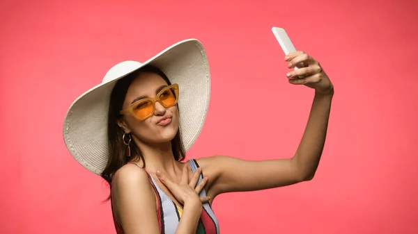 Mujer joven en sombrero de paja y traje de baño tomando selfie en el teléfono inteligente y mueca aislado en rosa - foto de stock