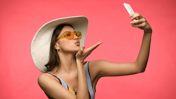 Молода модель в купальнику бере селфі на смартфон і дме повітряний поцілунок ізольовано на рожевому — стокове фото