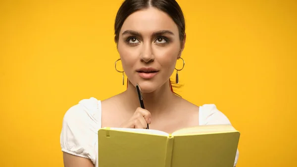 Pensive woman holding pen near notebook isolated on yellow — Stock Photo