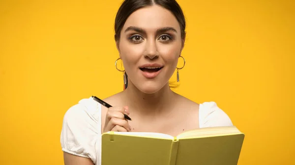 Amazed brunette woman holding pen near notebook isolated on yellow — стоковое фото