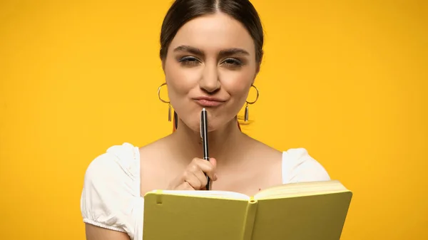 Sly brunette woman holding pen and looking at camera near notebook isolated on yellow — стоковое фото