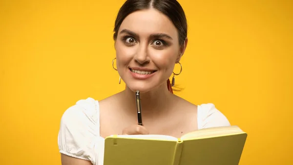 Brunette woman looking at camera while holding pen near notebook isolated on yellow - foto de stock