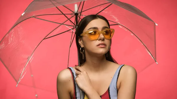 Молода жінка в сонцезахисних окулярах і купальнику тримає парасольку ізольовано на рожевому — стокове фото