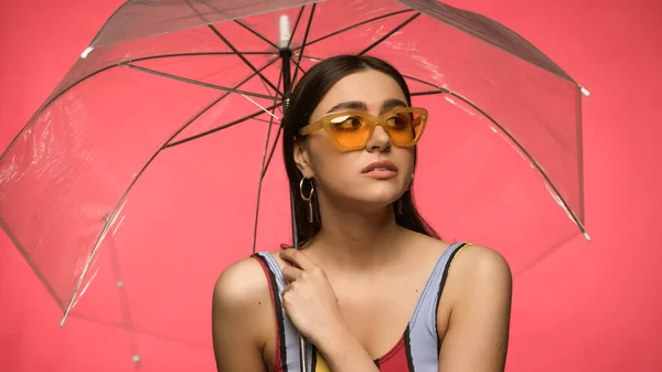 Young woman in sunglasses and swimsuit holding umbrella isolated on pink — Stockfoto