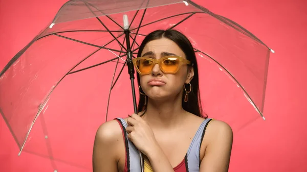 Displeased woman in swimsuit holding umbrella isolated on pink — стоковое фото