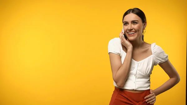 Smiling woman in blouse holding hand on hip and talking on smartphone isolated on yellow — Stock Photo