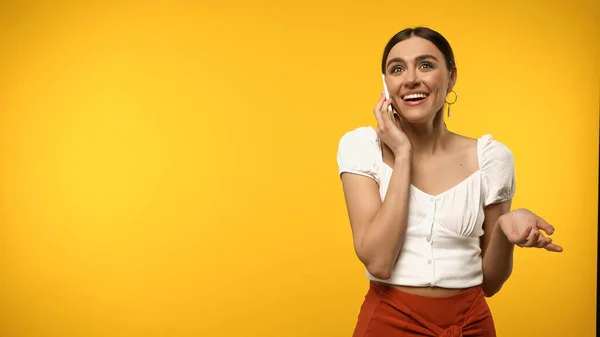 Positive brunette woman talking on smartphone and gesturing isolated on yellow — стоковое фото