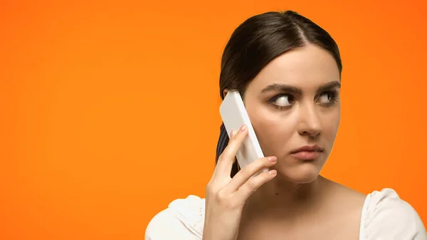 Modelo morena mirando hacia otro lado mientras habla en el teléfono inteligente aislado en naranja - foto de stock