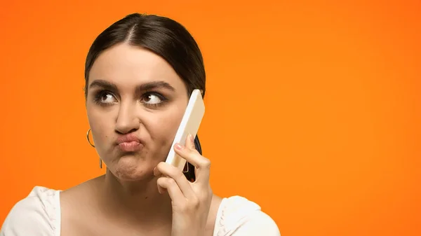 Femme brune confuse grimacant tout en parlant sur smartphone isolé sur orange — Photo de stock