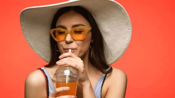 Pretty woman in sunglasses and swimsuit drinking cocktail isolated on coral - foto de stock