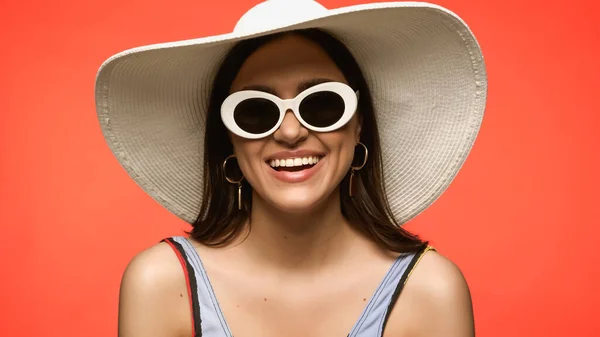 Mujer morena alegre en gafas de sol y sombrero de sol de pie aislado en coral - foto de stock