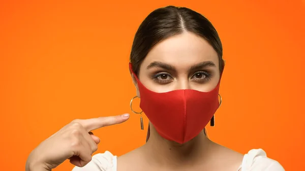 Brunette woman pointing with finger at protective mask isolated on orange — Stock Photo