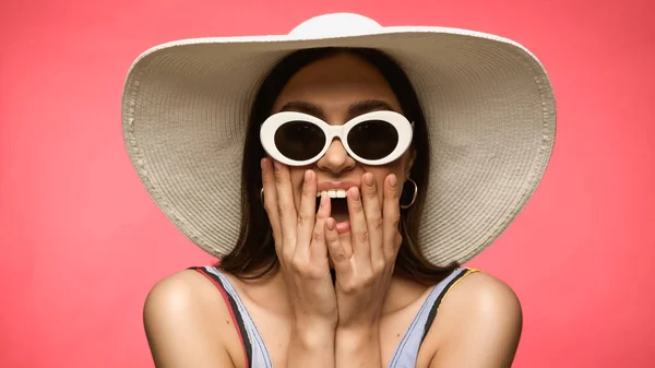 Дивовижна жінка в сонячному капелюсі і сонцезахисних окулярах торкається обличчя ізольовано на рожевому — стокове фото