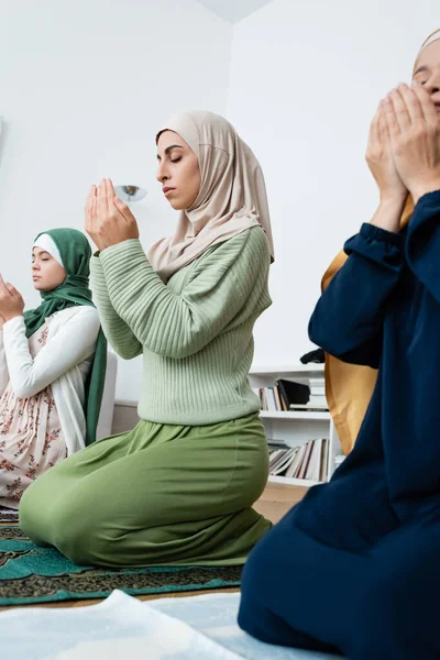 Junge muslimische Frau betet auf Teppich neben asiatischer Mutter und Tochter zu Hause — Stockfoto