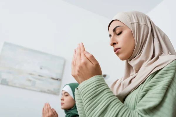Vista de ángulo bajo de la mujer árabe rezando cerca de hija borrosa en casa - foto de stock