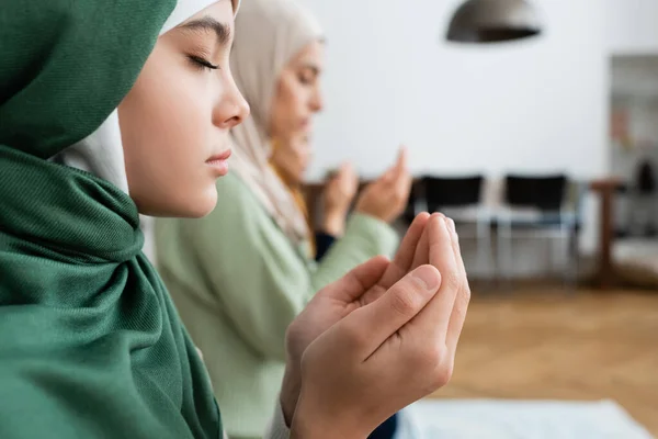 Vista lateral da menina no hijab orando perto da família turva em casa — Fotografia de Stock