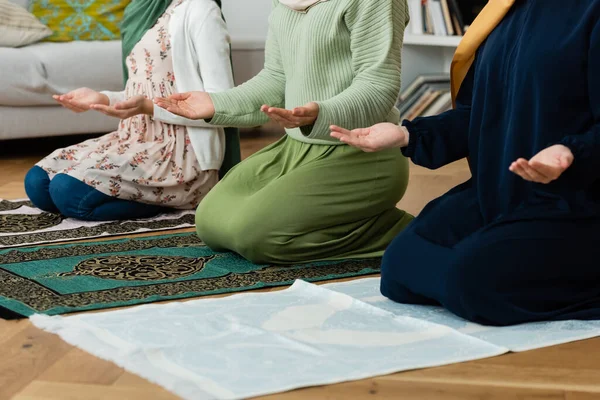 Cropped view of muslim women in traditional clothes praying on rugs at home — Stock Photo