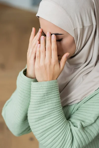 Mujer árabe joven cogida de la mano cerca de la cara mientras reza en casa - foto de stock