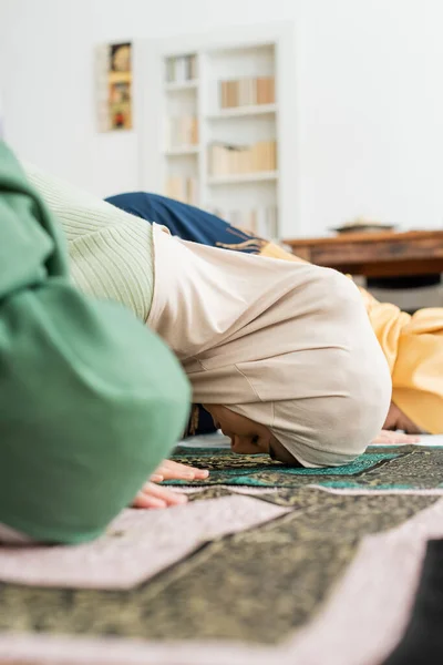 Vista lateral de la mujer árabe en hiyab tradicional rezando en trapo cerca de la familia en casa - foto de stock