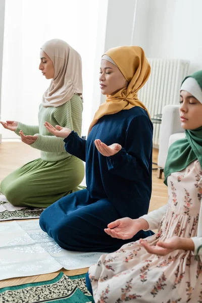 Femme asiatique en vêtements musulmans traditionnels priant près de fille arabe et petite-fille à la maison — Photo de stock