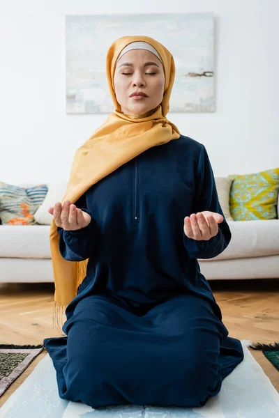 Asian woman praying with closed eyes on traditional rug at home — Stock Photo