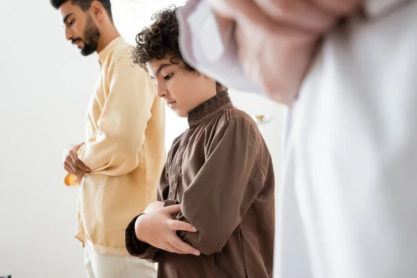 Muchacho musulmán rezando cerca de padre y borroso abuelo en casa - foto de stock
