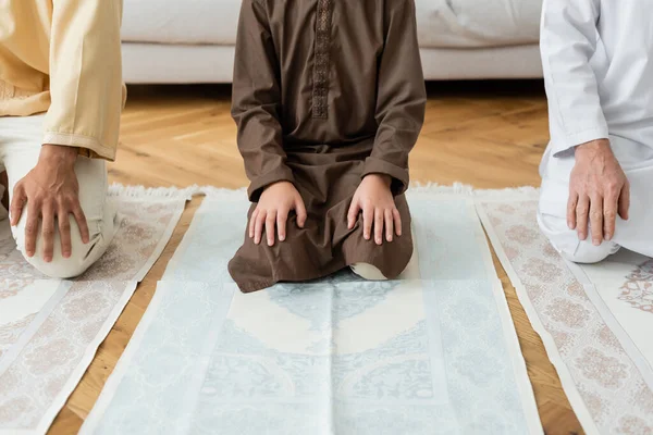 Vista recortada del niño musulmán preadolescente sentado en la alfombra cerca de la familia - foto de stock