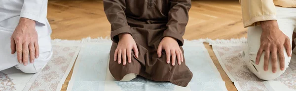 Vista ritagliata del bambino arabo seduto sul tappeto vicino alla famiglia a casa, striscione — Foto stock
