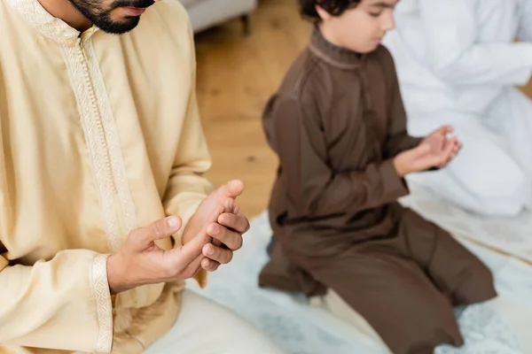 Hombre árabe joven rezando cerca de borrosa hijo y padre en casa - foto de stock