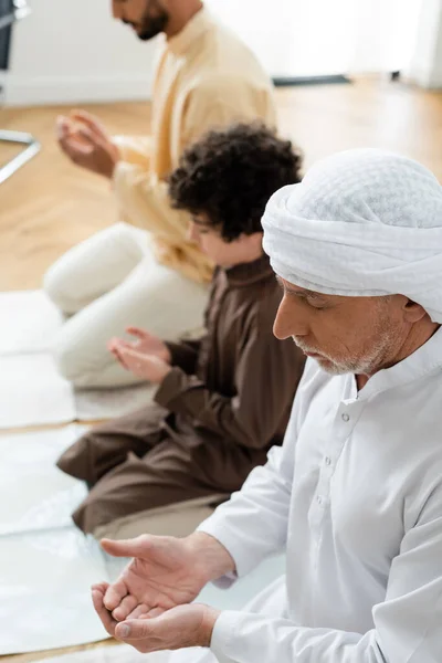 Hombre maduro rezando con los ojos cerrados cerca de hijo y niño musulmán en casa - foto de stock