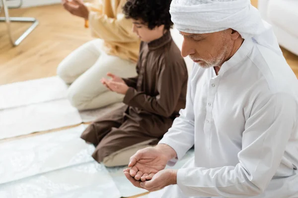 Mature man praying near blurred muslim grandson and son at home — Stock Photo